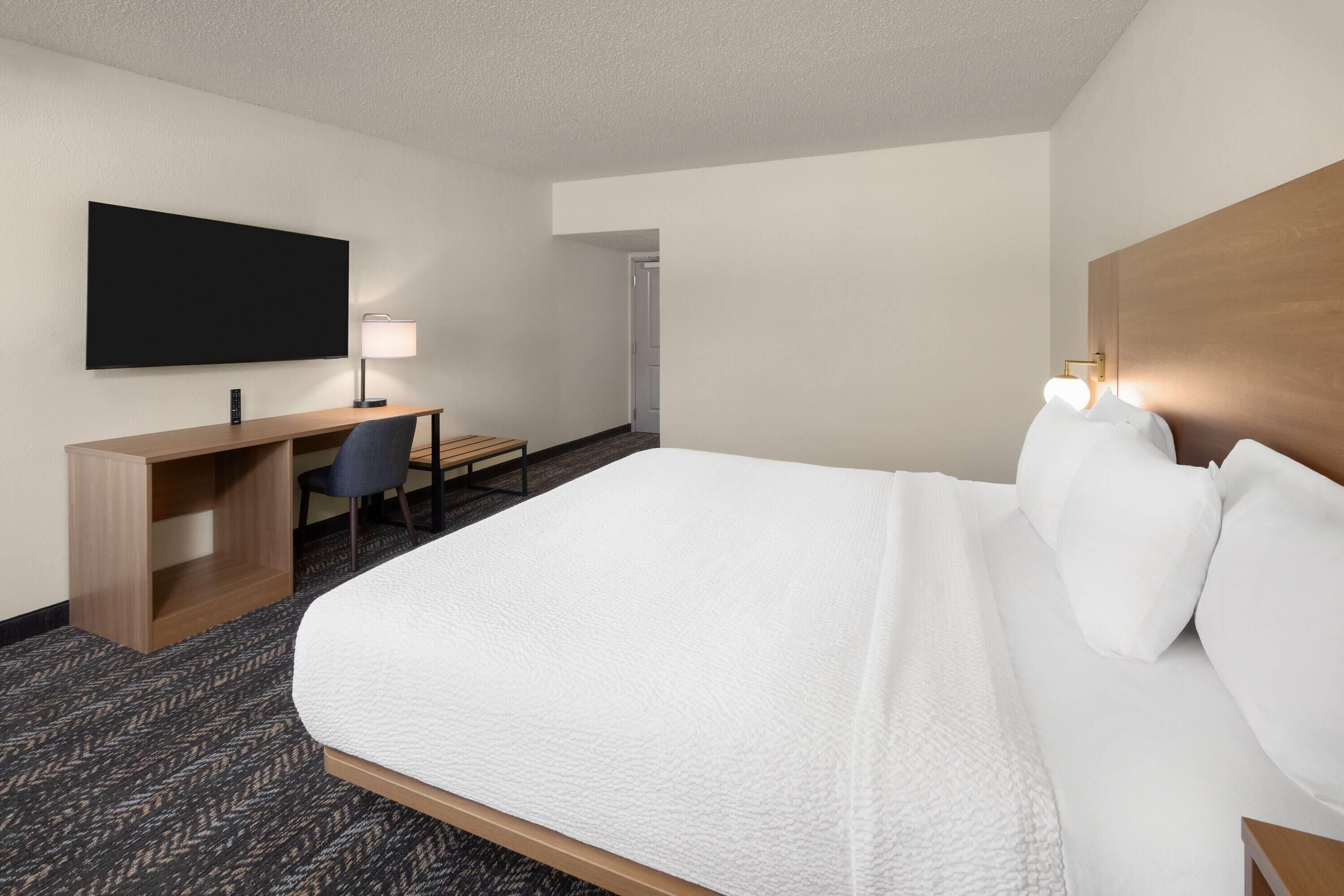 Quarto de hotel com uma cama grande, uma escrivaninha, uma cadeira, televisão de tela plana na parede e cabeceira de madeira.