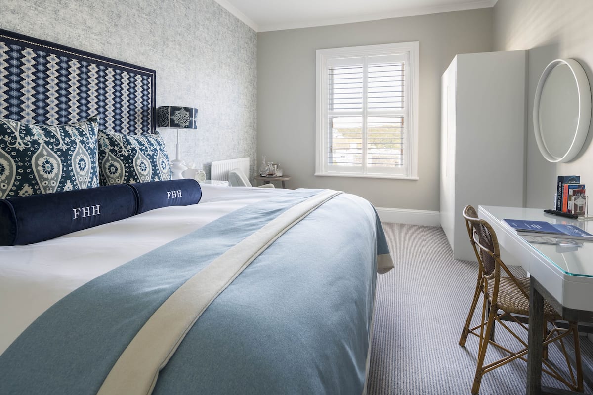 A hotel room with a large bed, a desk with a chair, a window with blinds, and a mirror.