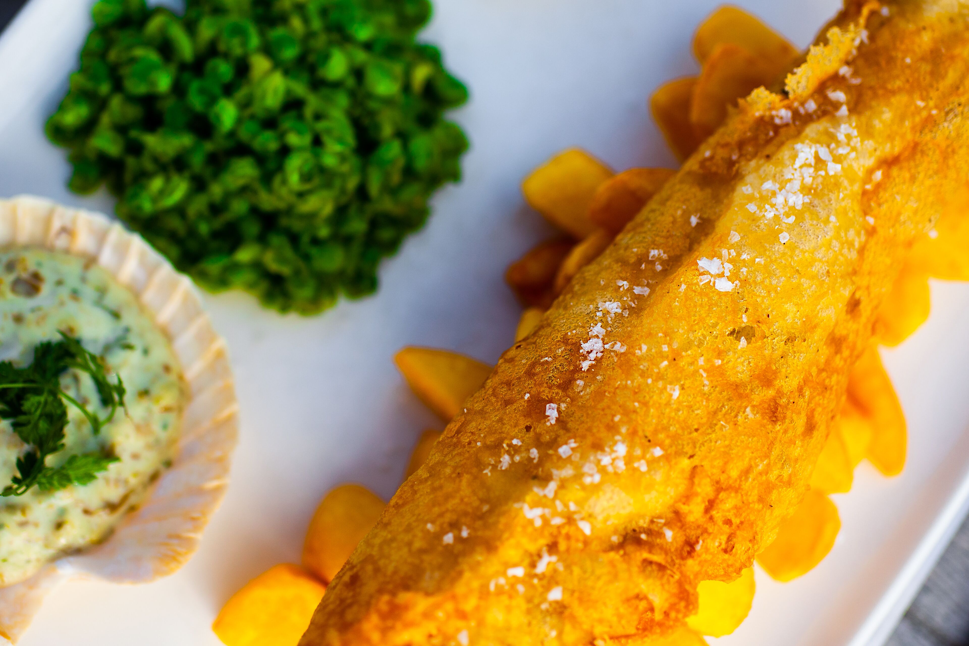 A plate with a large, golden-brown fried fish, a side of green peas, and a small bowl of creamy sauce.