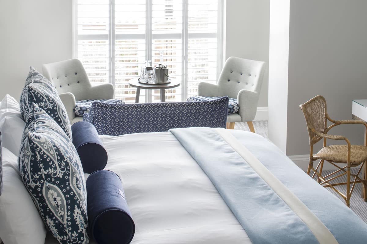 A modern bedroom with a large bed, two white armchairs, a small round table, and a wicker chair.