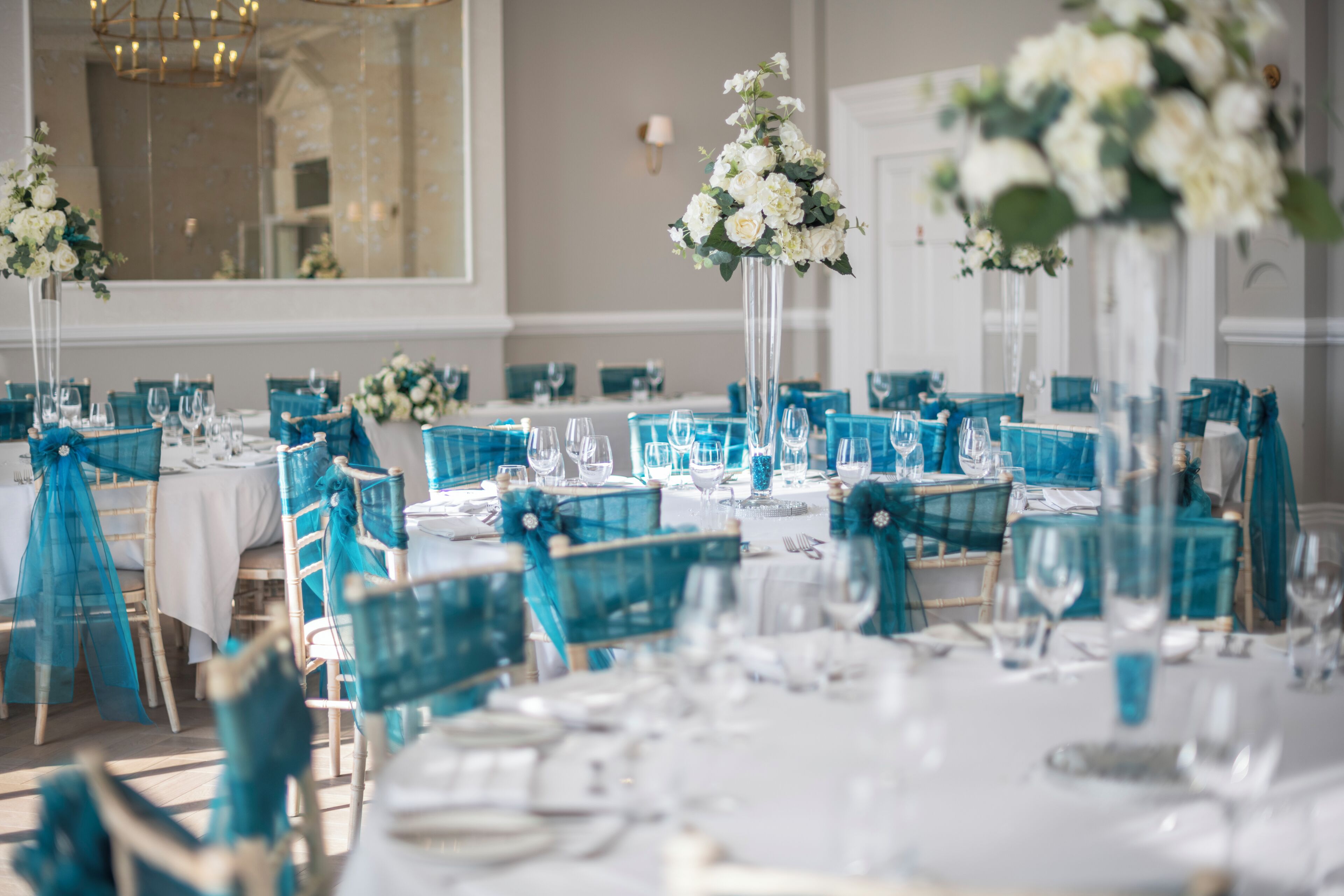 Salão de festas com mesas montadas para um evento formal, com cadeiras cor turquesa e toalhas de mesa brancas.