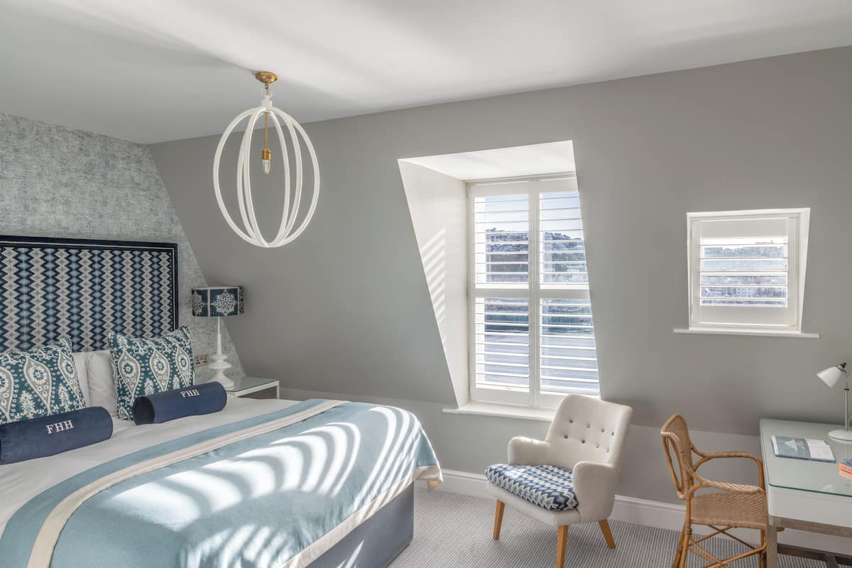 A bedroom with a large bed, a white chair, a desk, and a window with blinds.