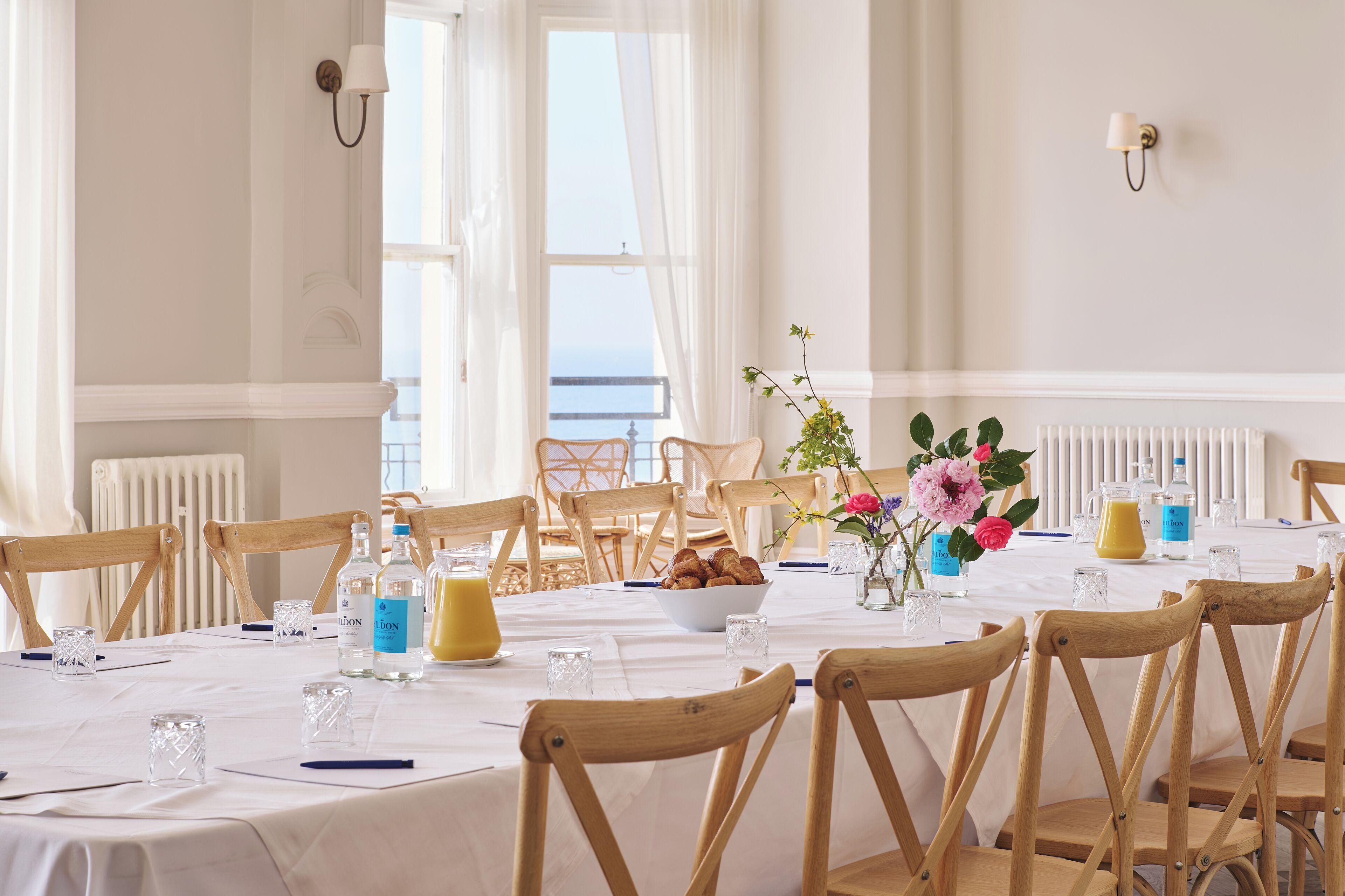A dining area with a white table set for six, wooden chairs, a vase with flowers, and a view of the sea through large windows.
