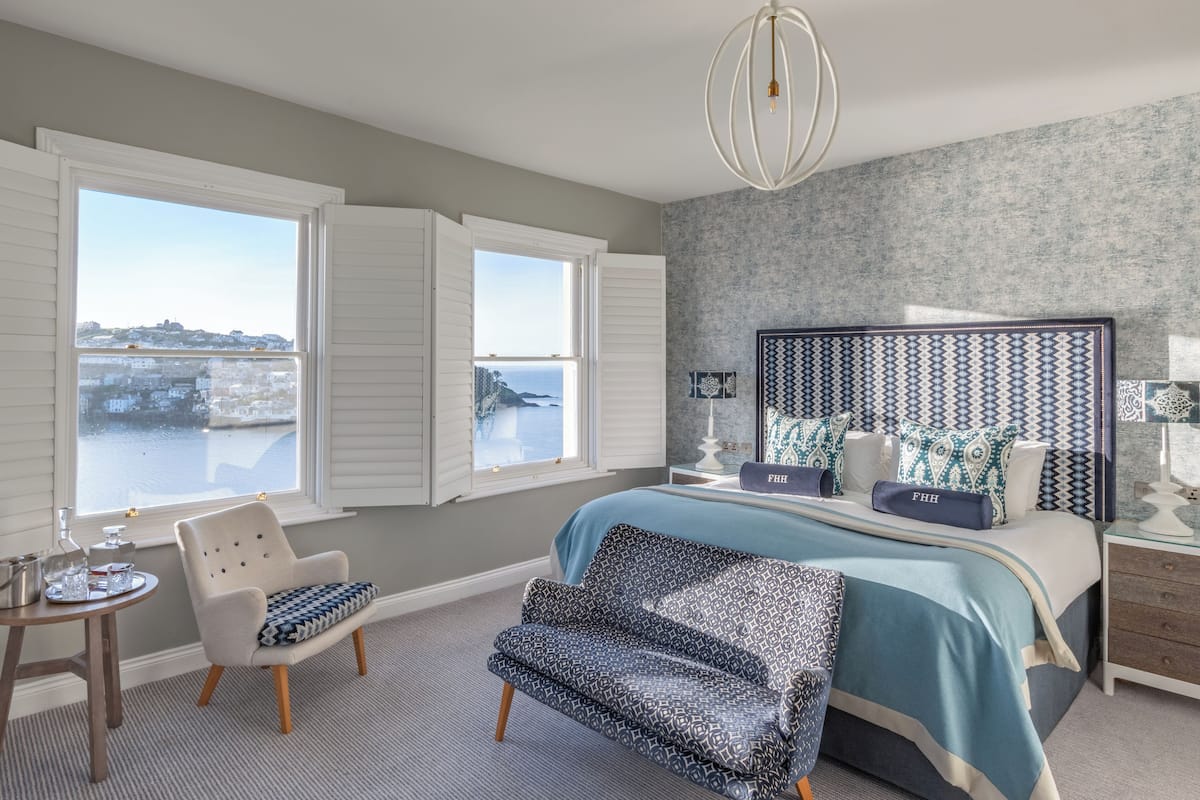 A bedroom with a large bed, a chair, a table with a glass top, and a view of the sea through open shutters.