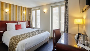 A hotel room with a large bed, a desk with a lamp, and a view of the city through the windows.