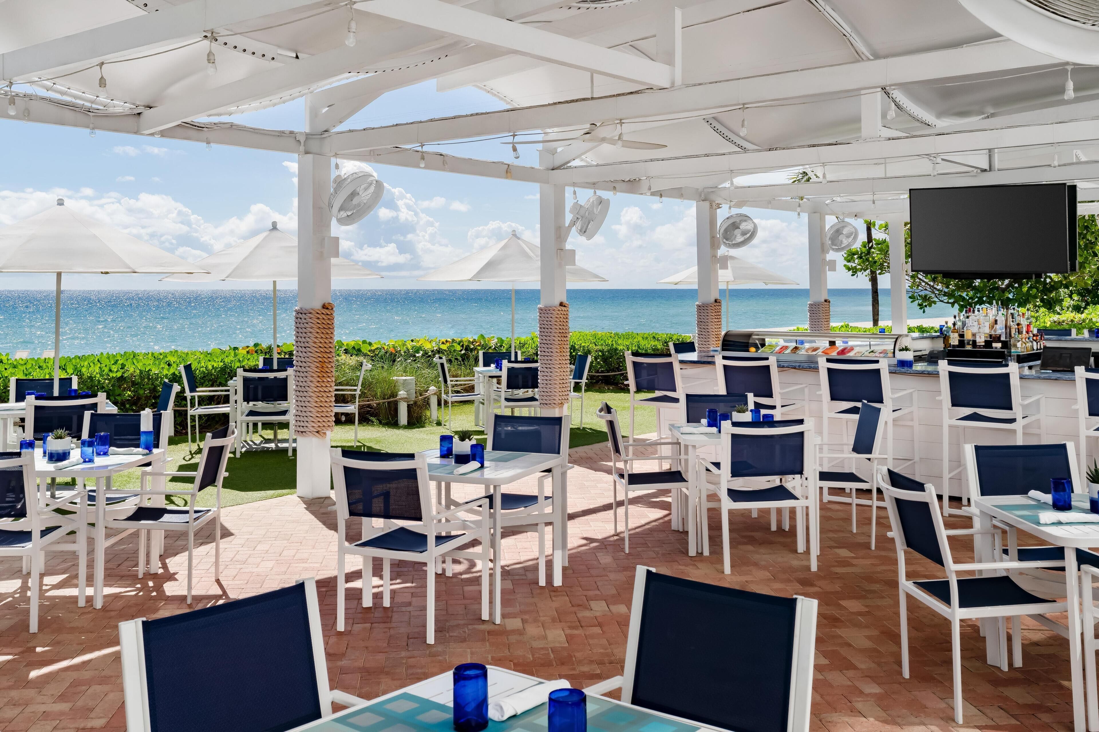 An outdoor dining area with white tables and chairs, a bar, and a view of the ocean.