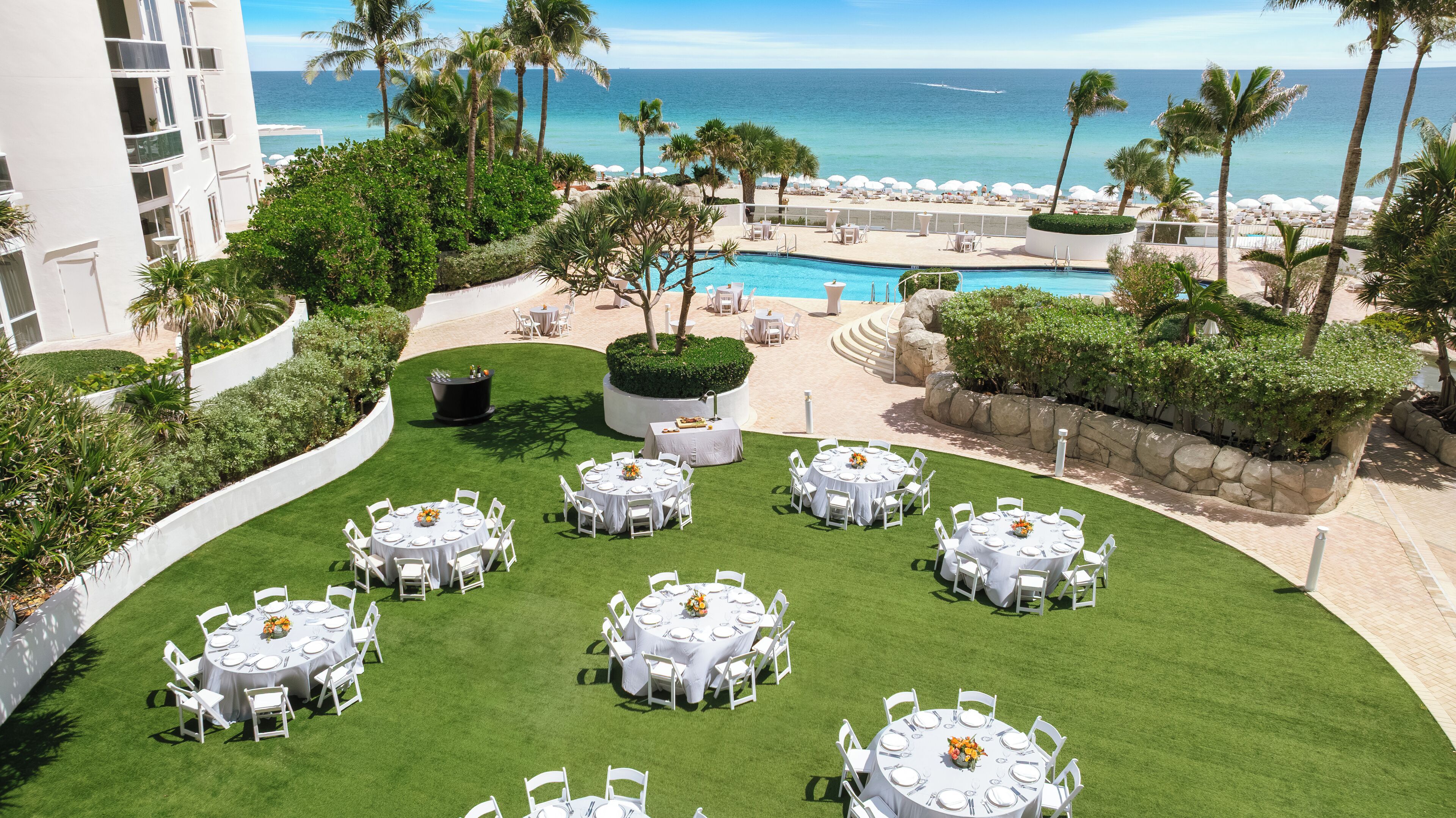 A resort with multiple round tables set up on a grassy lawn, overlooking a pool and the ocean.