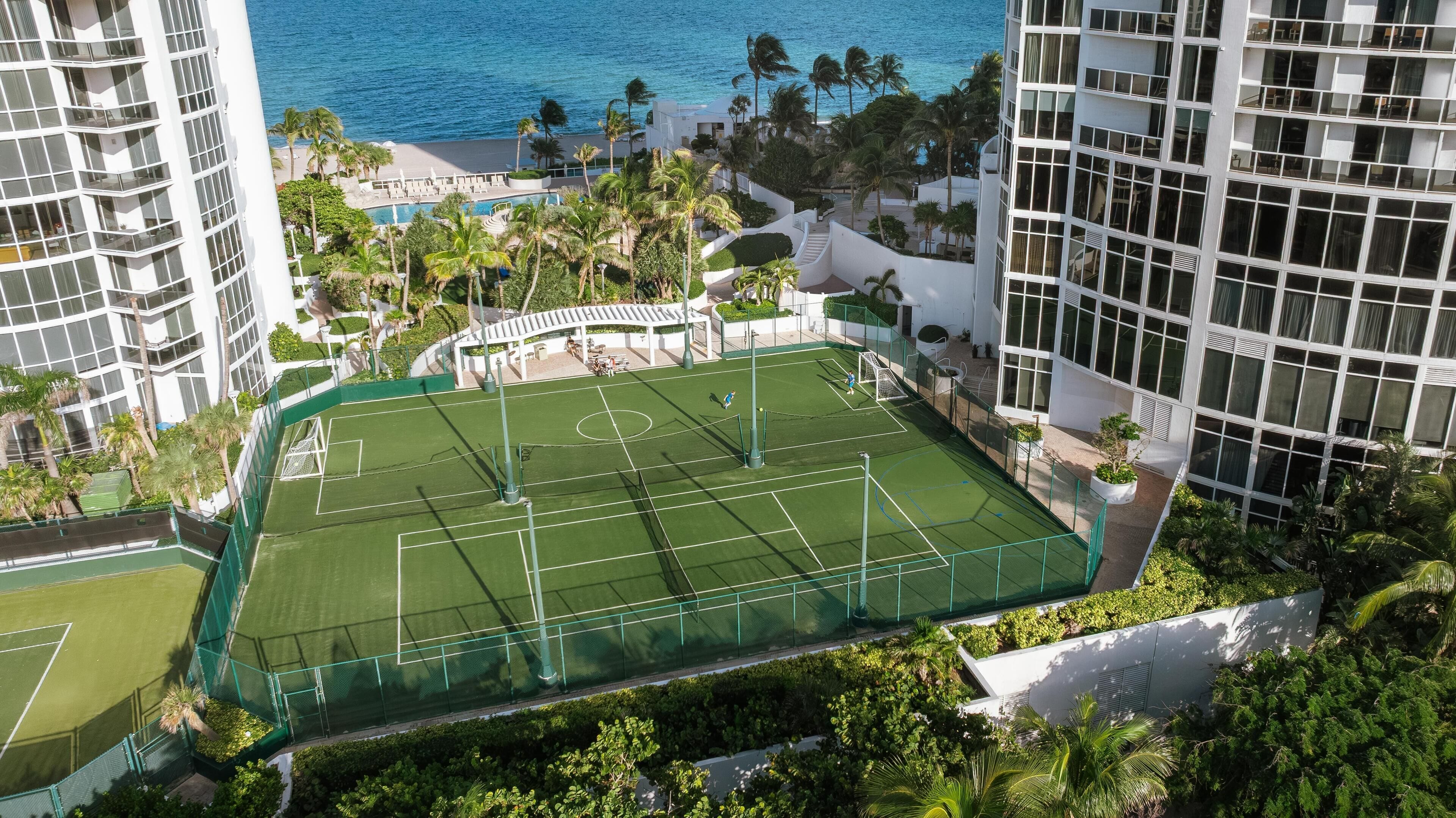 An aerial view of a tennis court surrounded by lush greenery and tall buildings, with a pool and palm trees nearby.