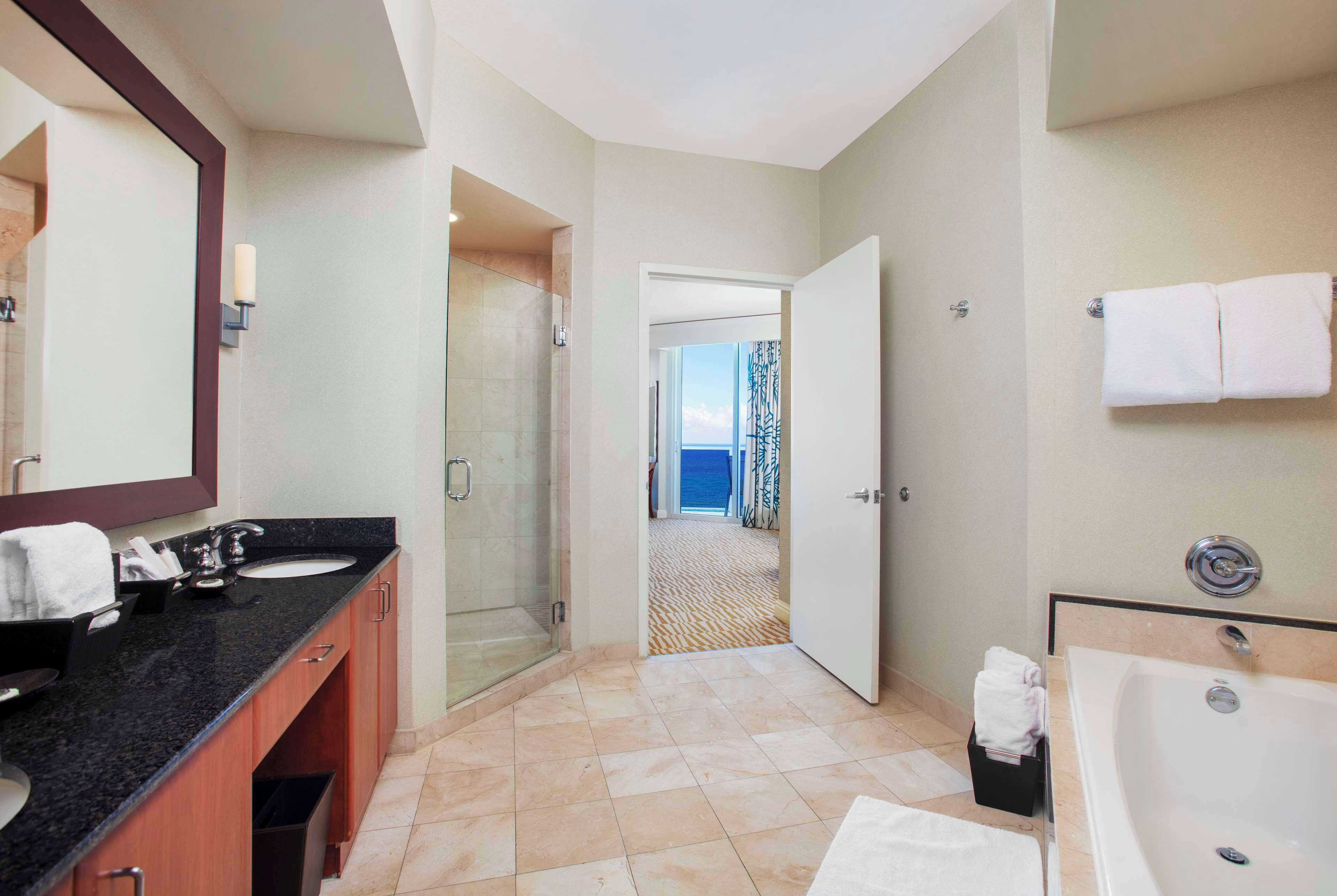 A hotel bathroom with a large bathtub, a separate shower area, a vanity with a mirror, and a view through an open door.