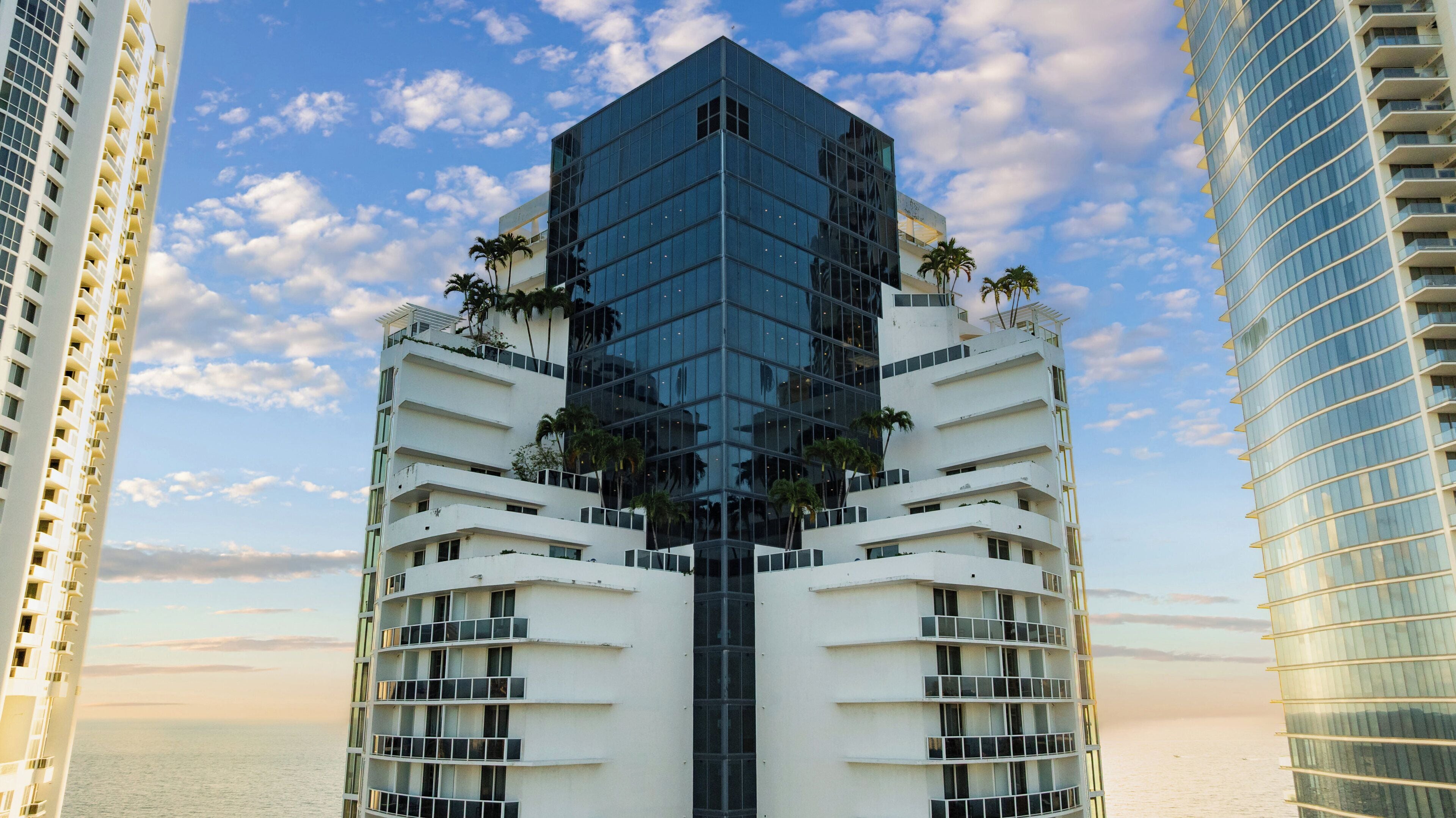 A modern high-rise building with a glass facade, surrounded by palm trees and a clear sky.