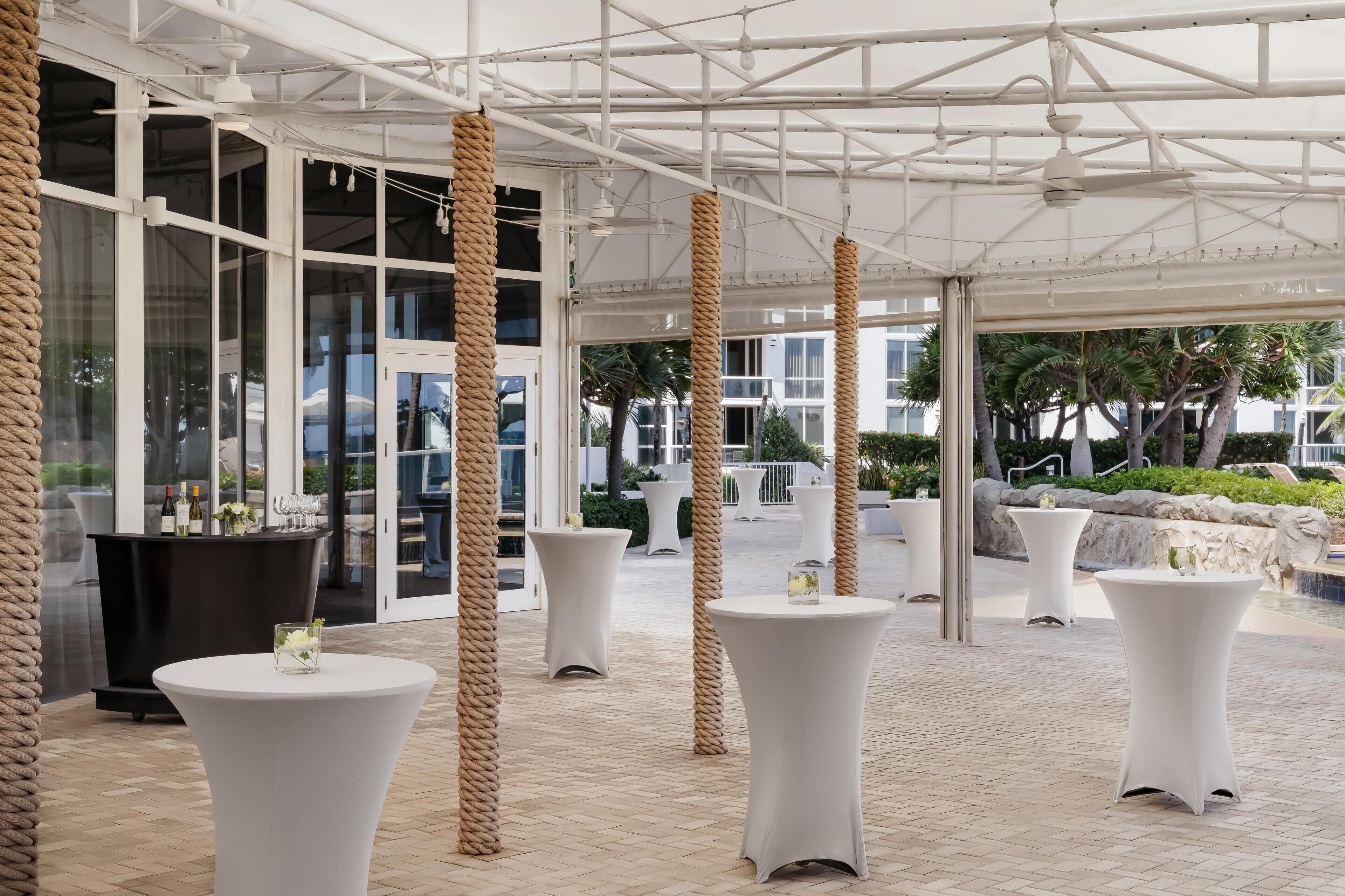 A covered outdoor area with white tables and chairs, a bar area with bottles, and a view of the outdoors.
