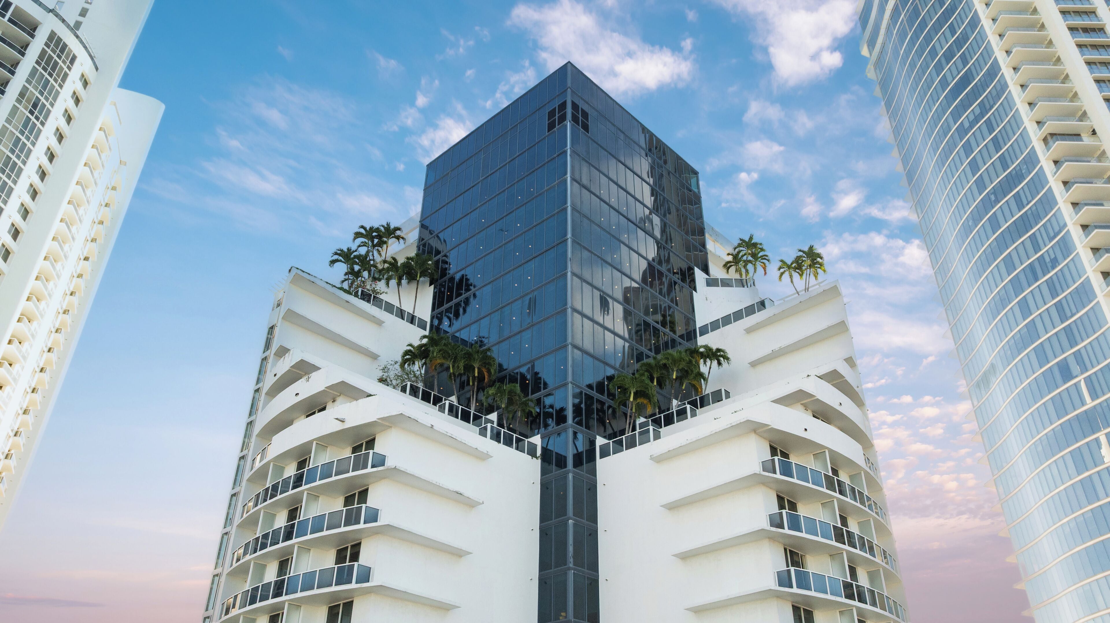 Un moderno edificio de gran altura con fachada de vidrio, rodeado de palmeras y otros edificios de gran altura.