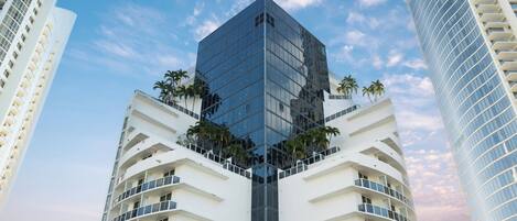 A modern high-rise building with a glass facade, surrounded by palm trees and other high-rise buildings.