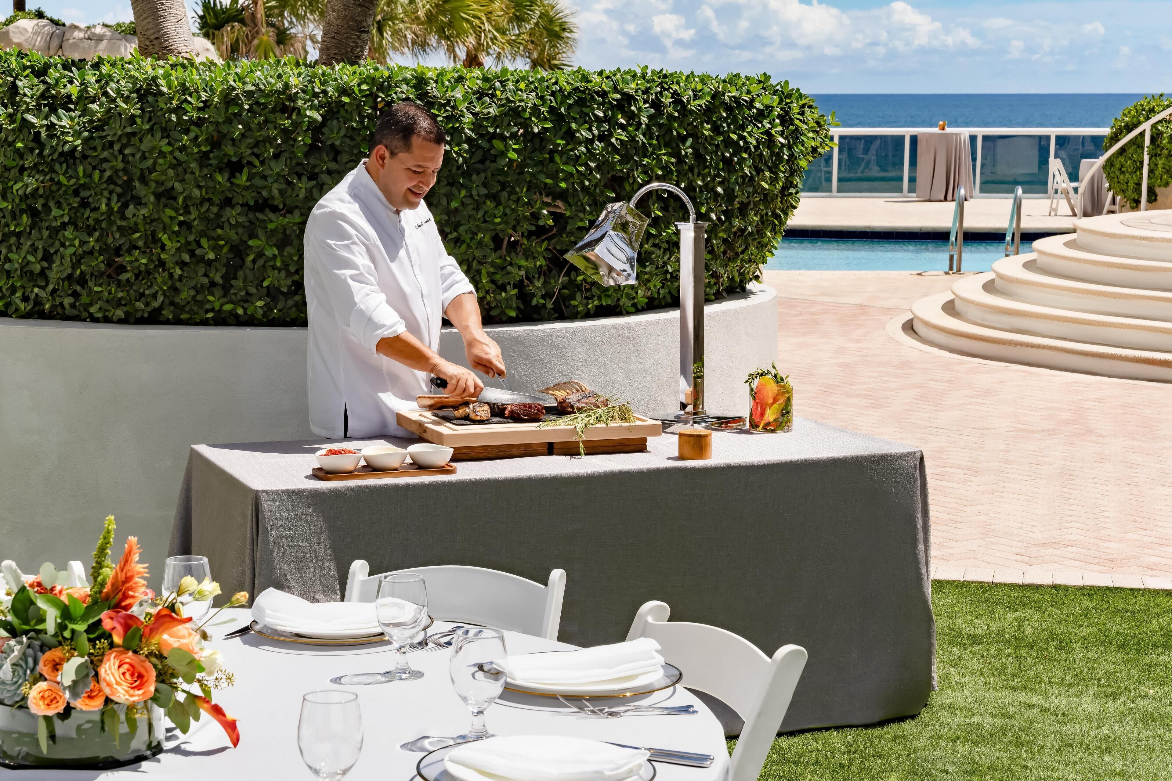 Uno chef prepara del cibo a un tavolo all'aperto con vista sull'oceano.