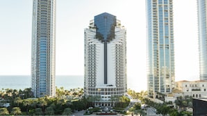 A high-rise hotel with a central tower, surrounded by palm trees and a view of the sea.