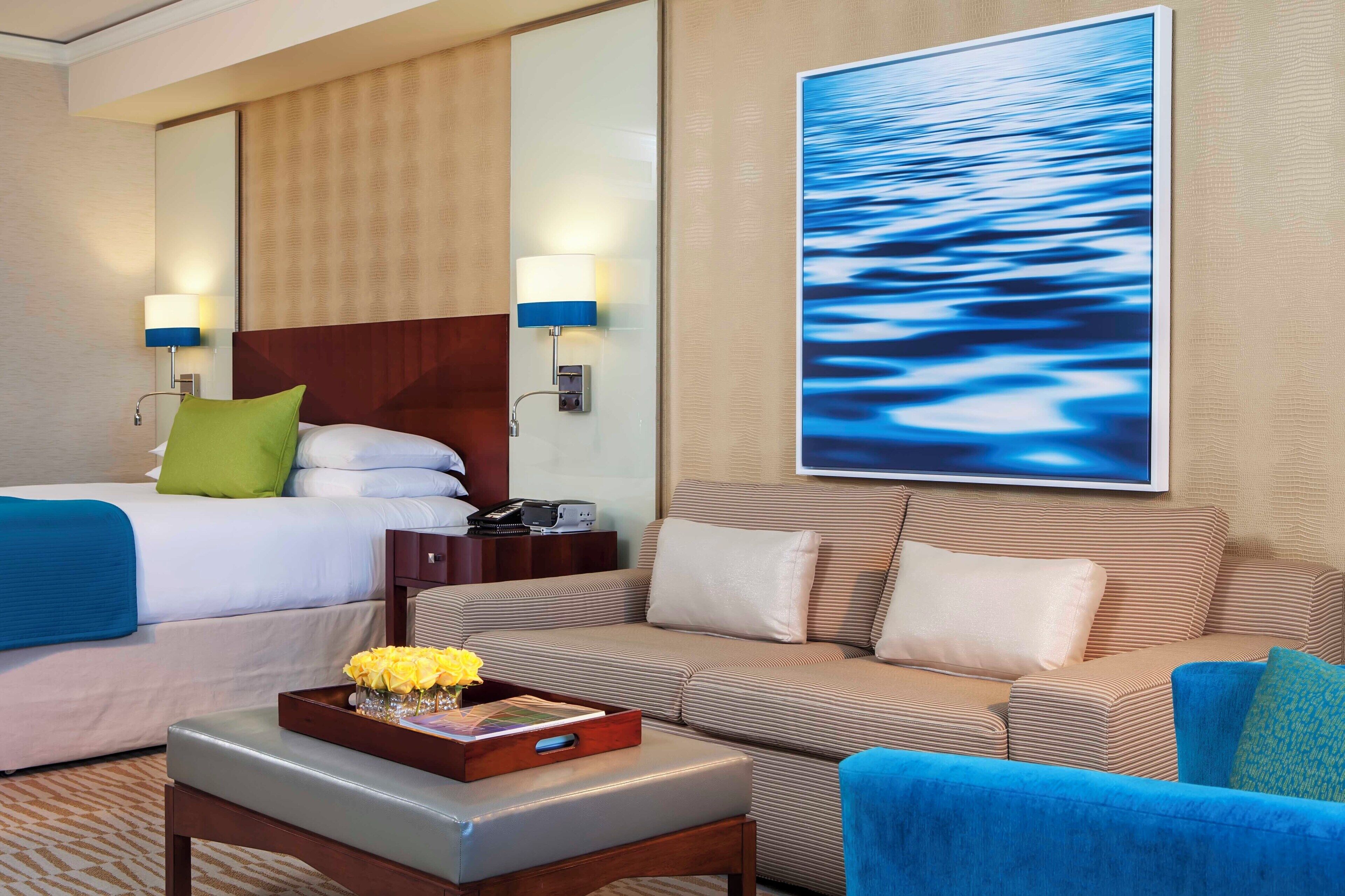 A hotel room with a bed, a sofa, a coffee table with a tray, and a large painting of water on the wall.