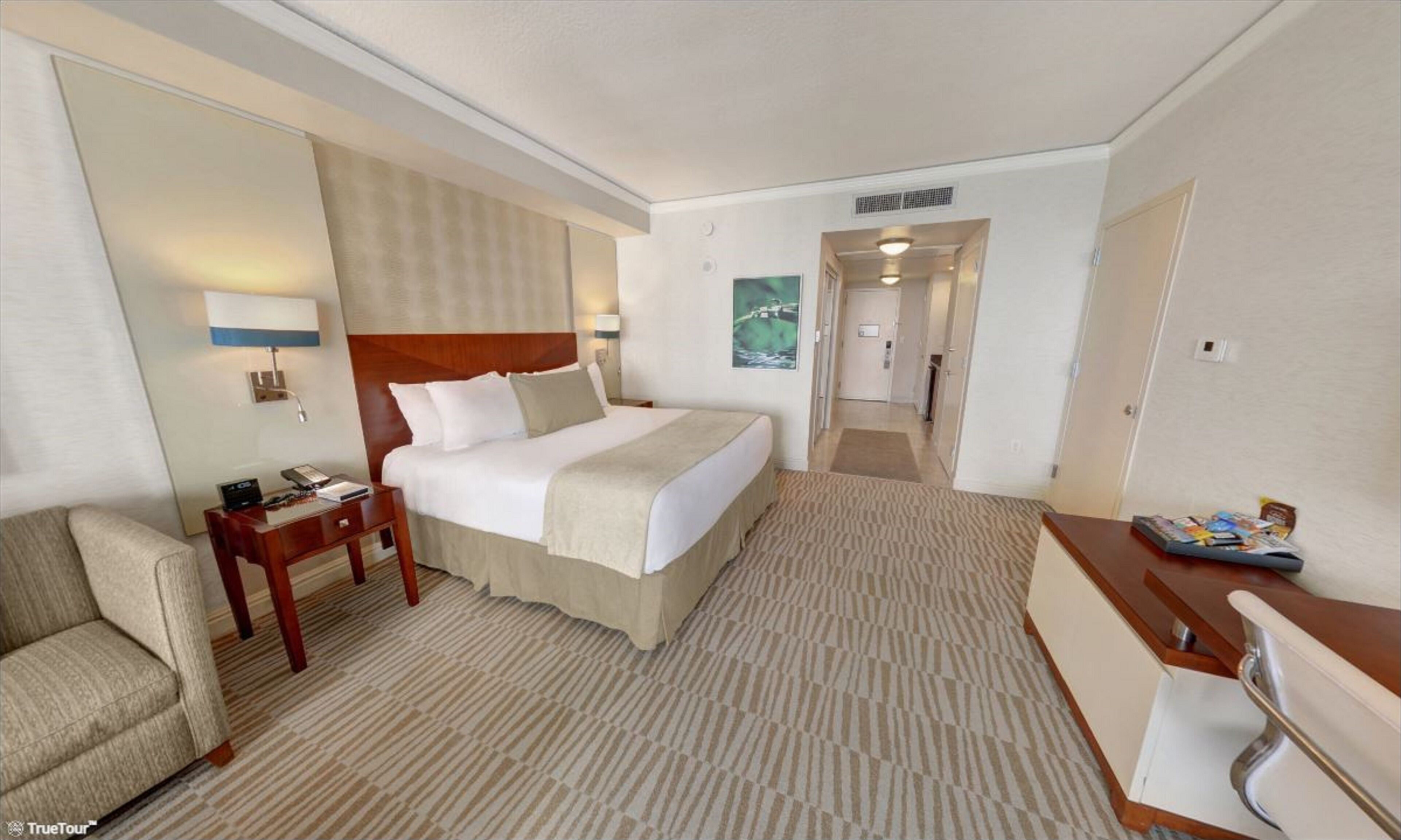 A hotel room with a bed, bedside table, lamp, chair, desk, and a door leading to another room.