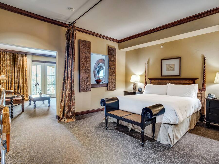 Een hotelkamer met een groot bed, een zitbankje, een bureau met een lamp en uitzicht op het omliggende landschap door een raam.