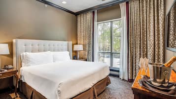 A hotel room with a large bed, a nightstand, a bedside table with a lamp, a window with curtains, and a table with a champagne bucket.
