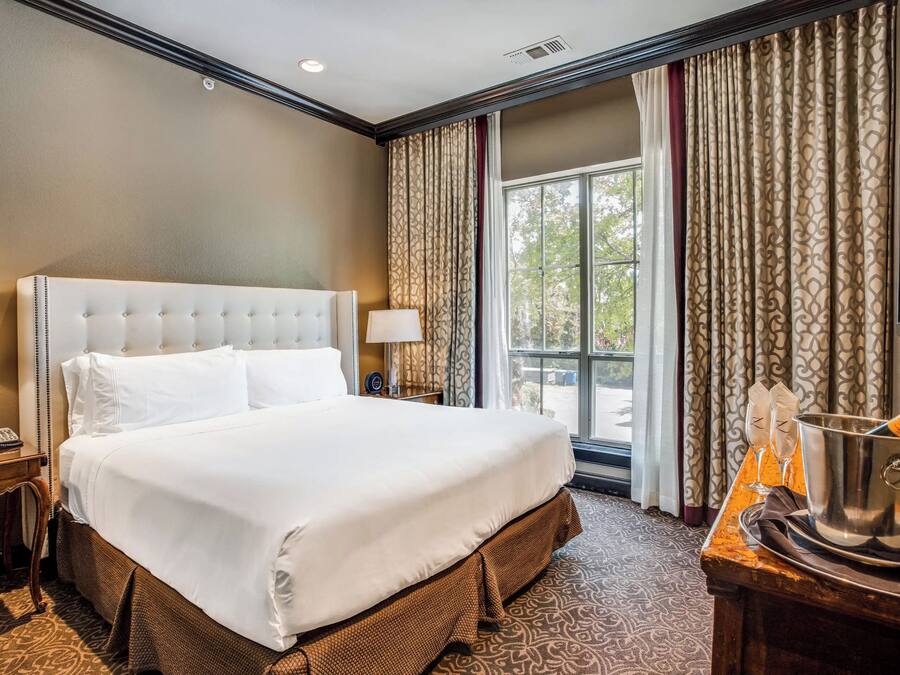 A hotel room with a large bed, a nightstand, a bedside table with a lamp, a window with curtains, and a table with a champagne bucket.