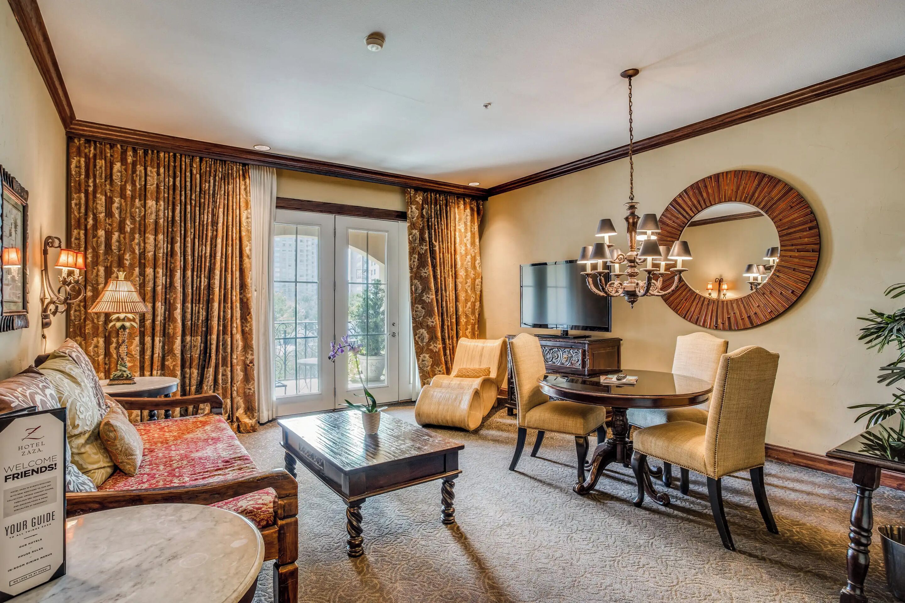 A hotel room with a dining area, a chandelier, a large mirror, and a view through the window.