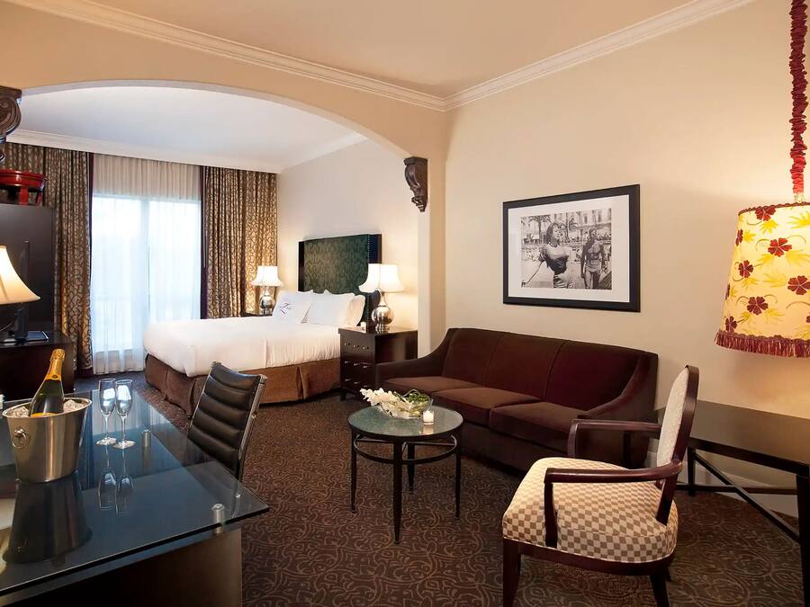 Habitación de hotel con cama, sofá, mesa de comedor con una botella de champán, dos copas de vino, una mesita con una vela y un cuadro en la pared.