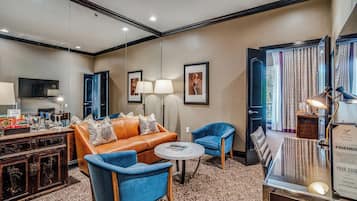 Habitación de hotel con sofá de cuero, sillones azules, una mesa de centro redonda, televisor y escritorio con lámpara.