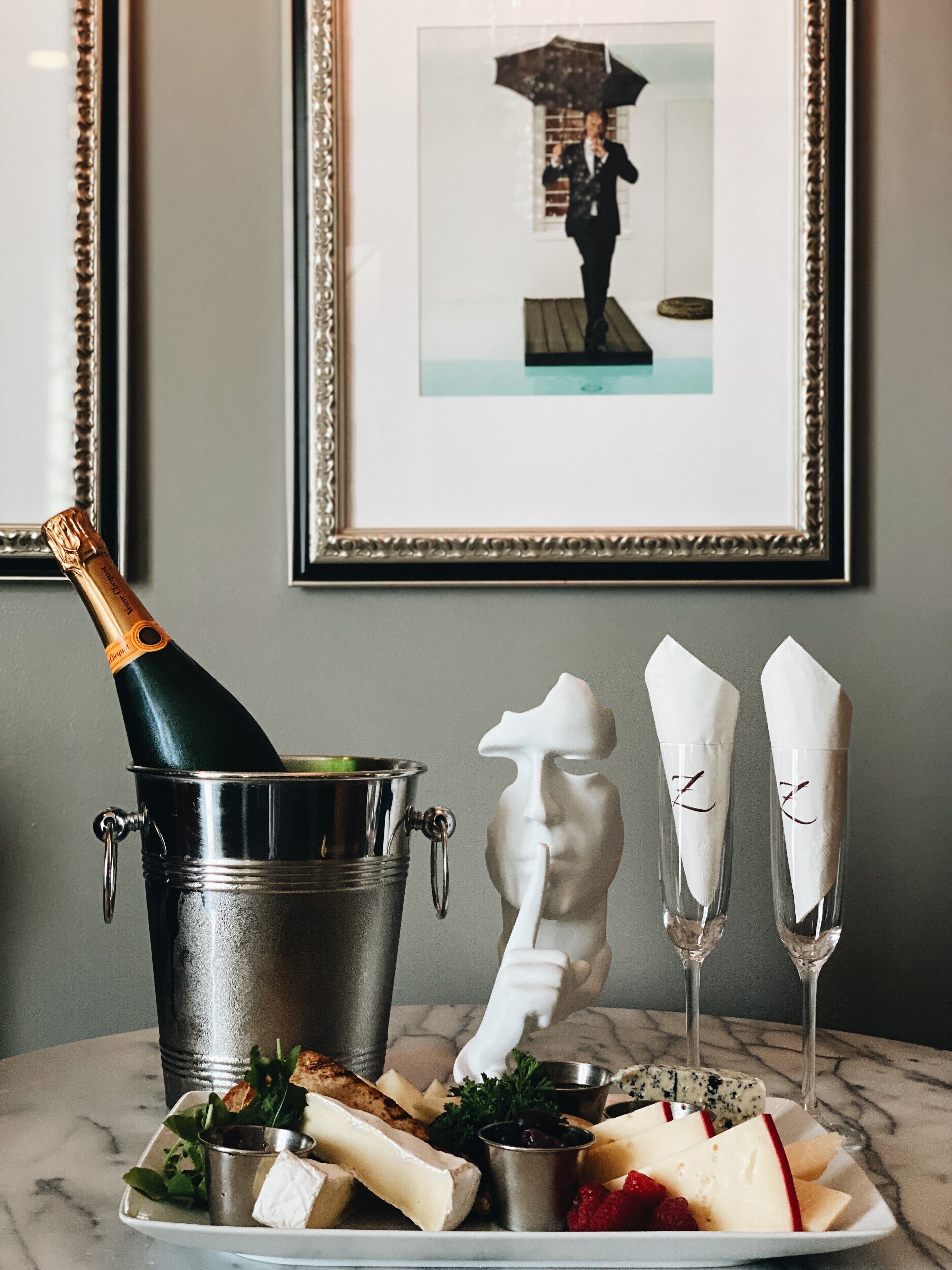 A table with a bottle of champagne, a white sculpture, two glasses, and a plate of cheese and fruits.