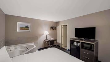 A hotel room with a large bed, a desk with a lamp, a TV, a minibar, and a bathroom with a bathtub.