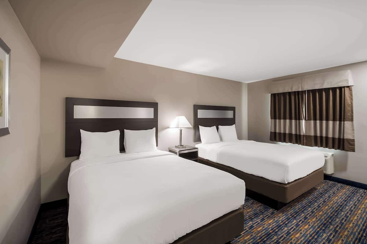 Habitación de hotel con dos camas, una mesita de noche con lámpara, una ventana con cortinas y piso alfombrado.