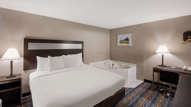A hotel room with a large bed, a desk, a chair, a desk lamp, a bedside table, a nightstand, a wall-mounted TV, and a bathroom with a bathtub and a shower. - Americas Best Value Inn Farmington (Farmington)
