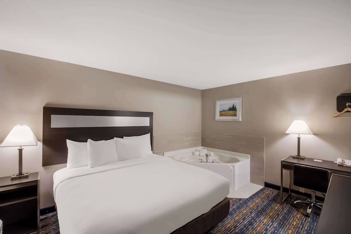 Habitación de hotel con una cama grande, un escritorio, una silla, una lámpara de escritorio, una mesita de noche, televisor montado en la pared y un baño con bañera y ducha.