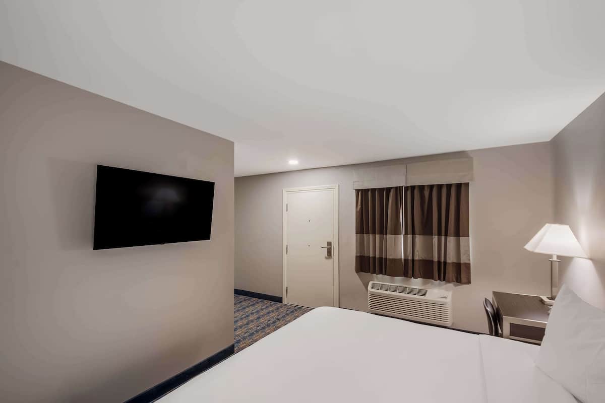 Habitación de hotel con cama, escritorio con silla, televisor montado en la pared, puerta y ventana con cortinas.