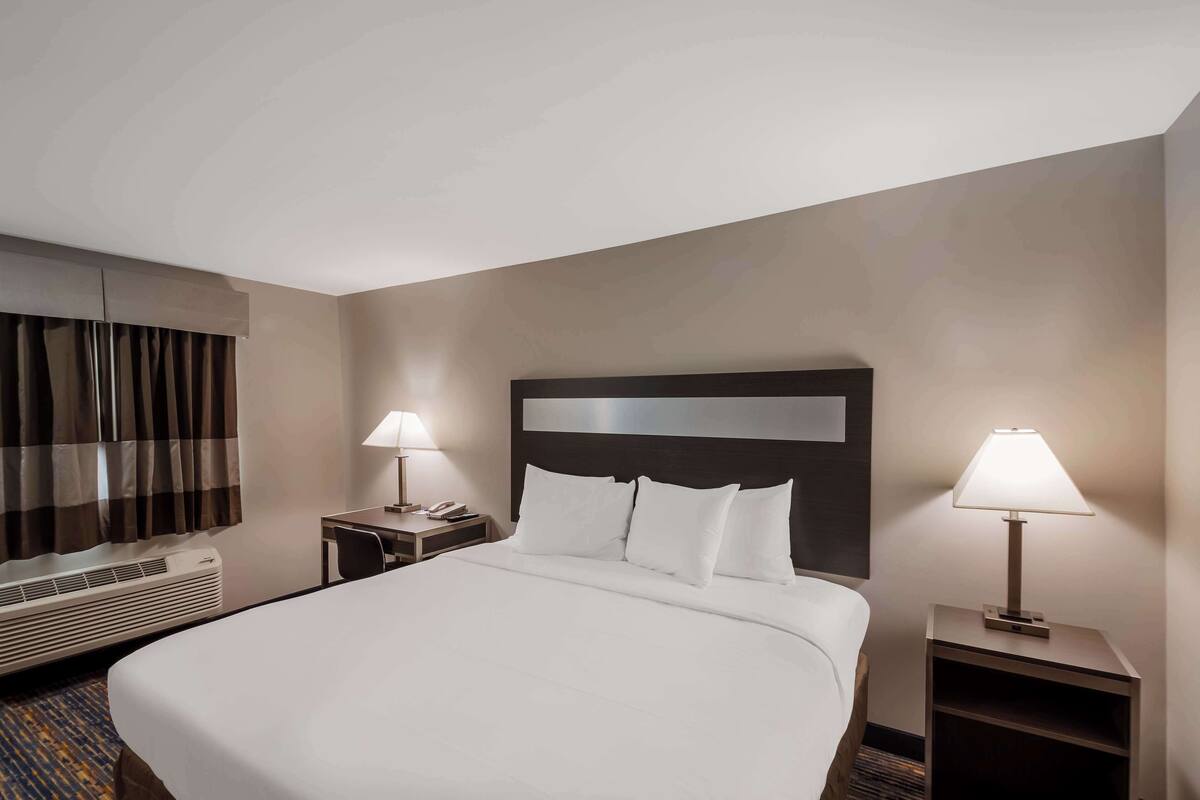 A hotel room with a large bed, two bedside tables with lamps, a headboard, an air conditioning unit, and a window with curtains.
