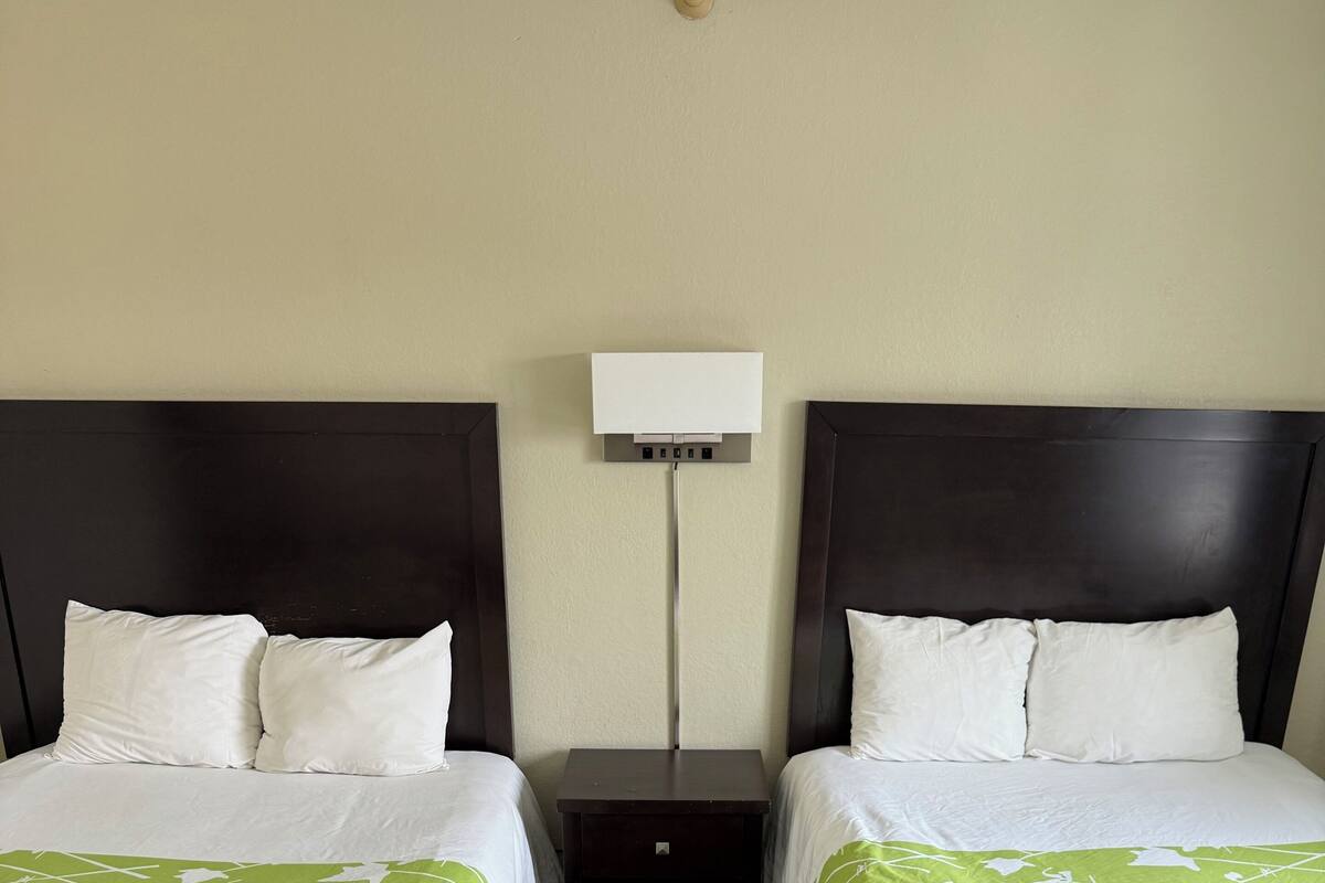 Hotelkamer met twee bedden, elk voorzien van wit beddengoed en groene accenten, een donker houten hoofdbord, een nachtkastje met een lamp en een houten vloer.