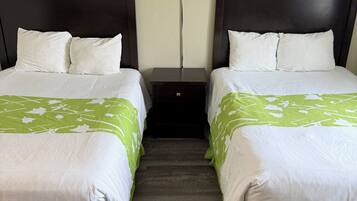 A hotel room with two beds, each with white bedding and green accents, a dark wooden headboard, a nightstand with a lamp, and a wooden floor.