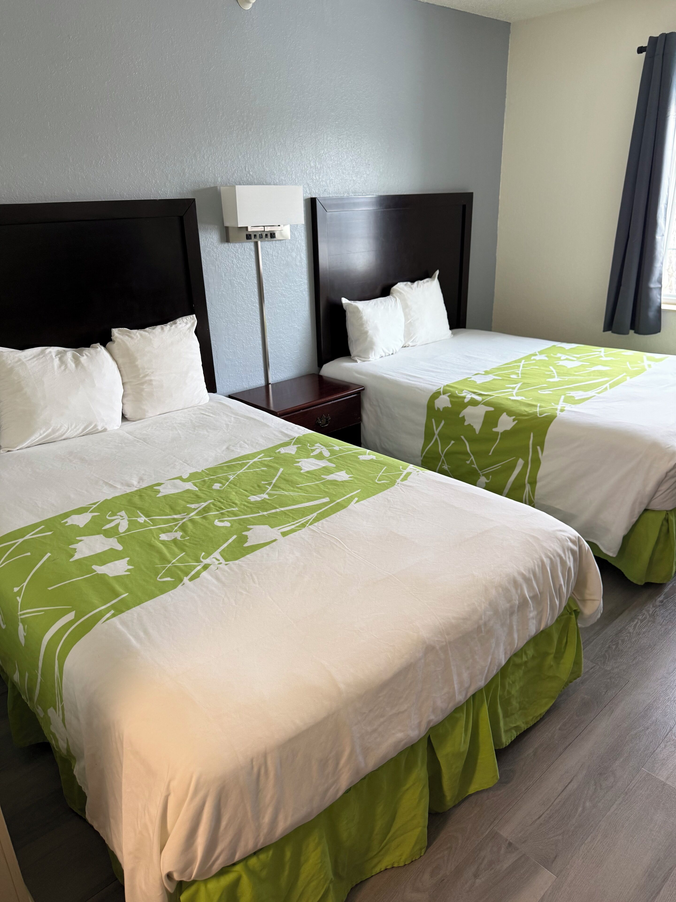 Dos camas con ropa de cama blanca y verde, una mesita de noche, una lámpara y una ventana con cortinas.