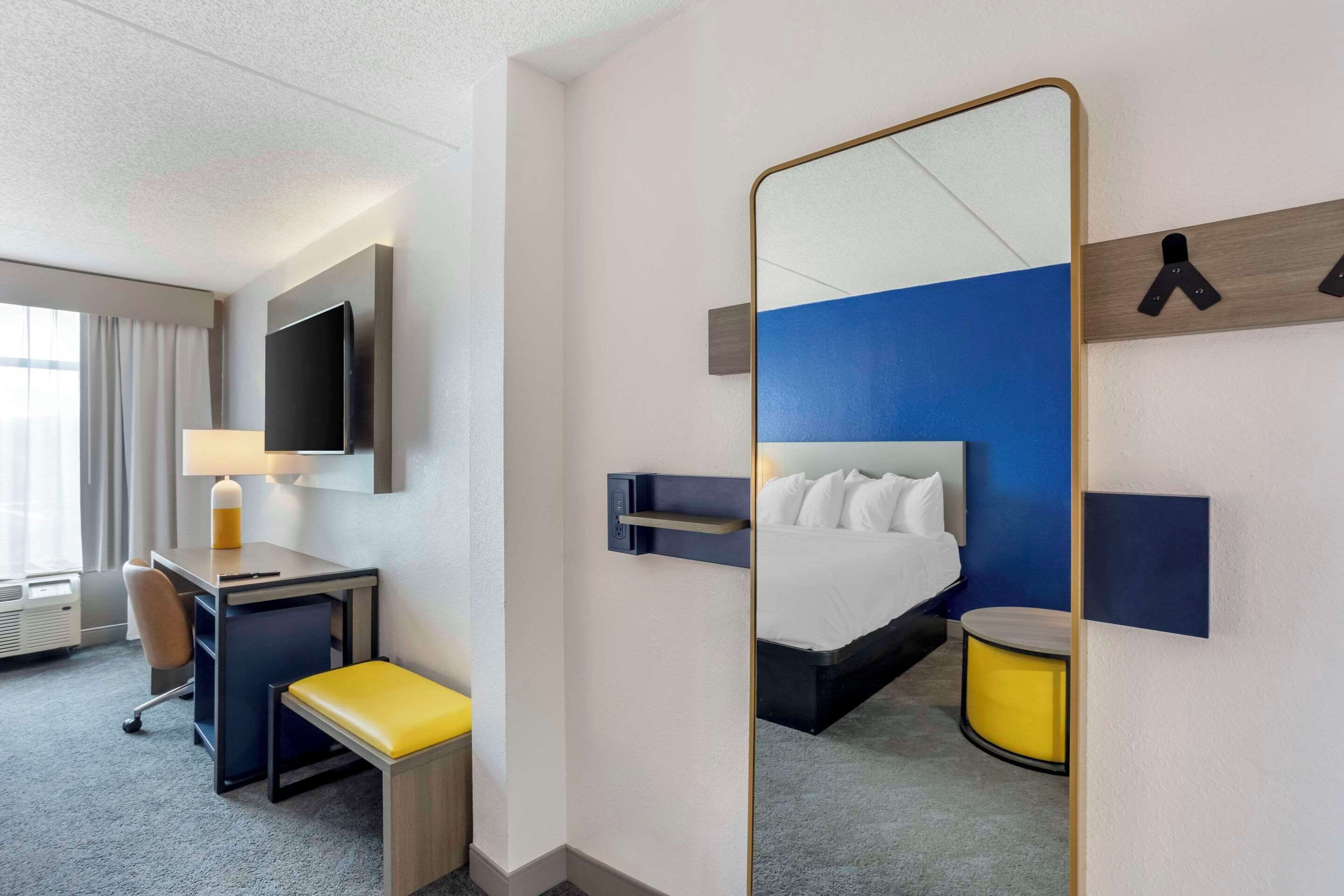 A hotel room with a bed, a desk with a chair, a small table, a mirror, and a wall-mounted TV.
