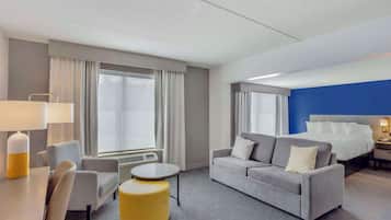 A hotel room with a bed, a sofa, a small table, and a chair. There is a window with curtains, a lamp, and a heater.