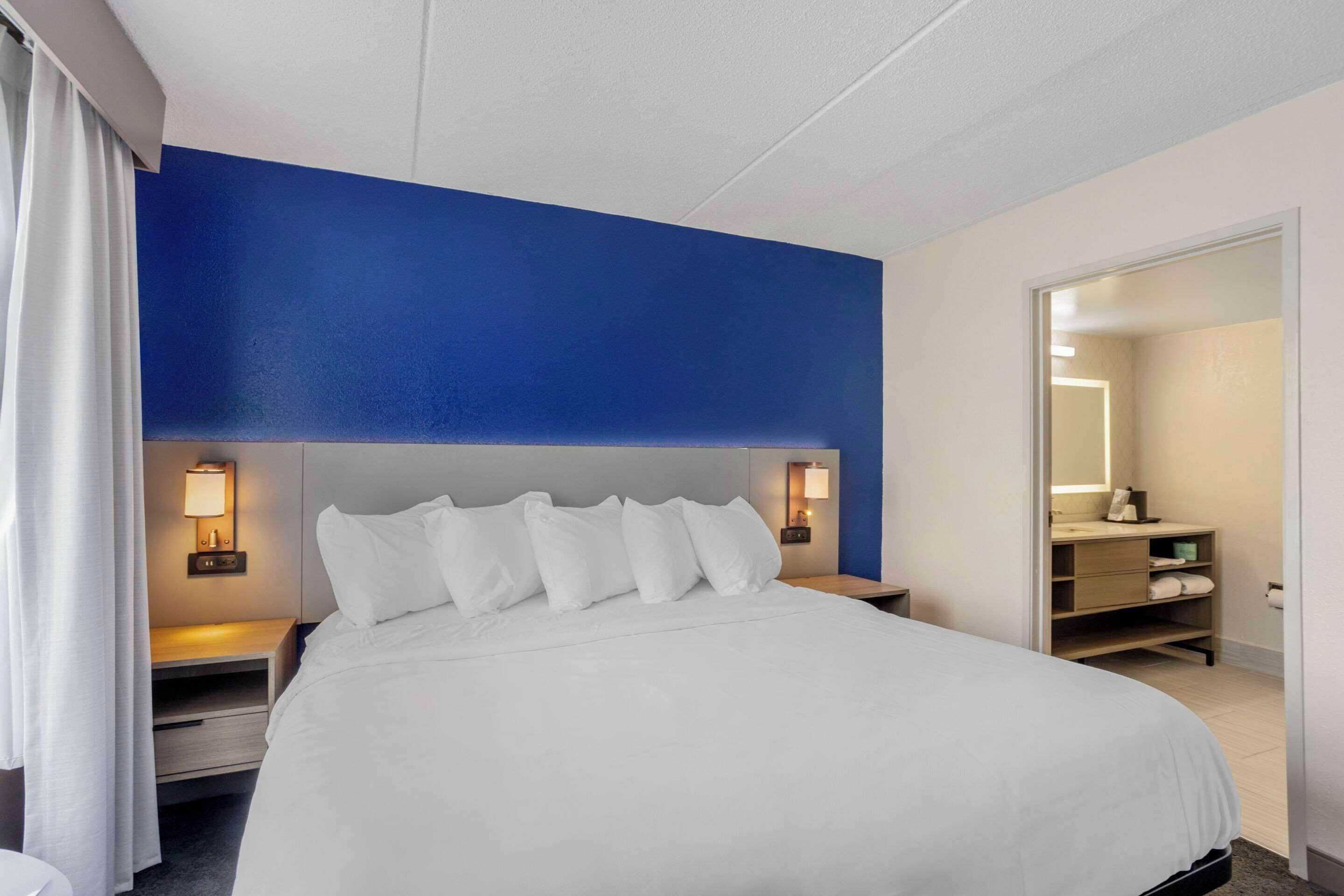 A hotel room with a large bed, two bedside tables with lamps, a blue accent wall, and a bathroom visible through an open door.