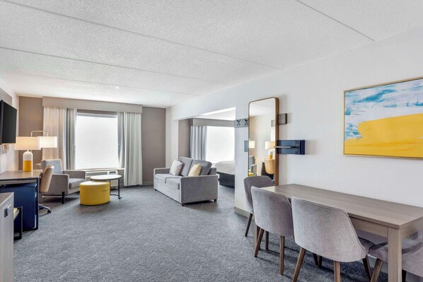A hotel room with a grey sofa, a small round table, a desk with two chairs, a TV, and a large window with curtains. - Comfort Inn & Suites Voorhees/Mt. Laurel (Voorhees)