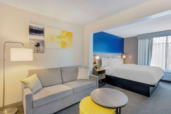A hotel room with a bed, a sofa, a bedside table, a lamp, and artwork on the wall.