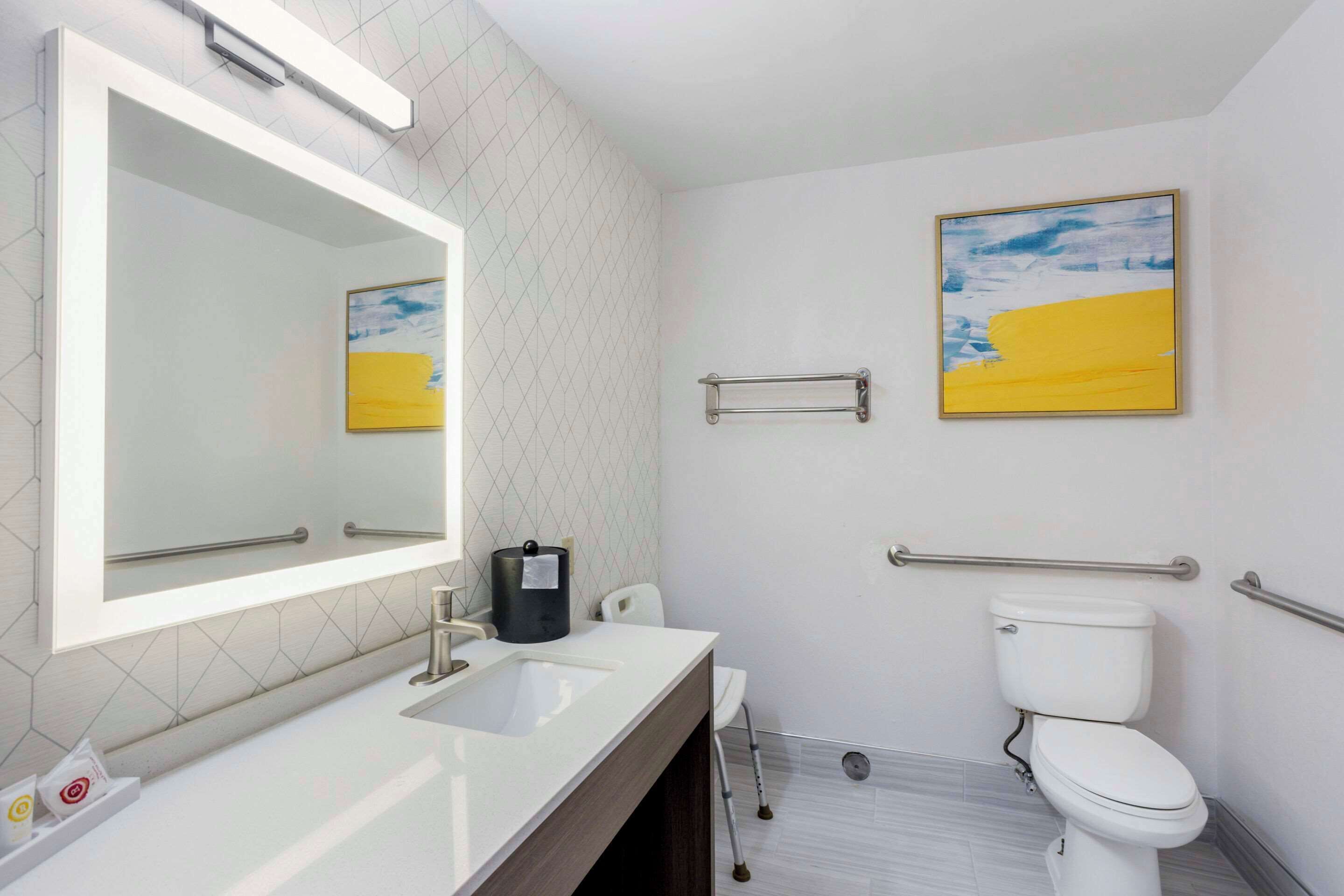 A modern bathroom with a large mirror, a white sink, a toilet, and a handrail.