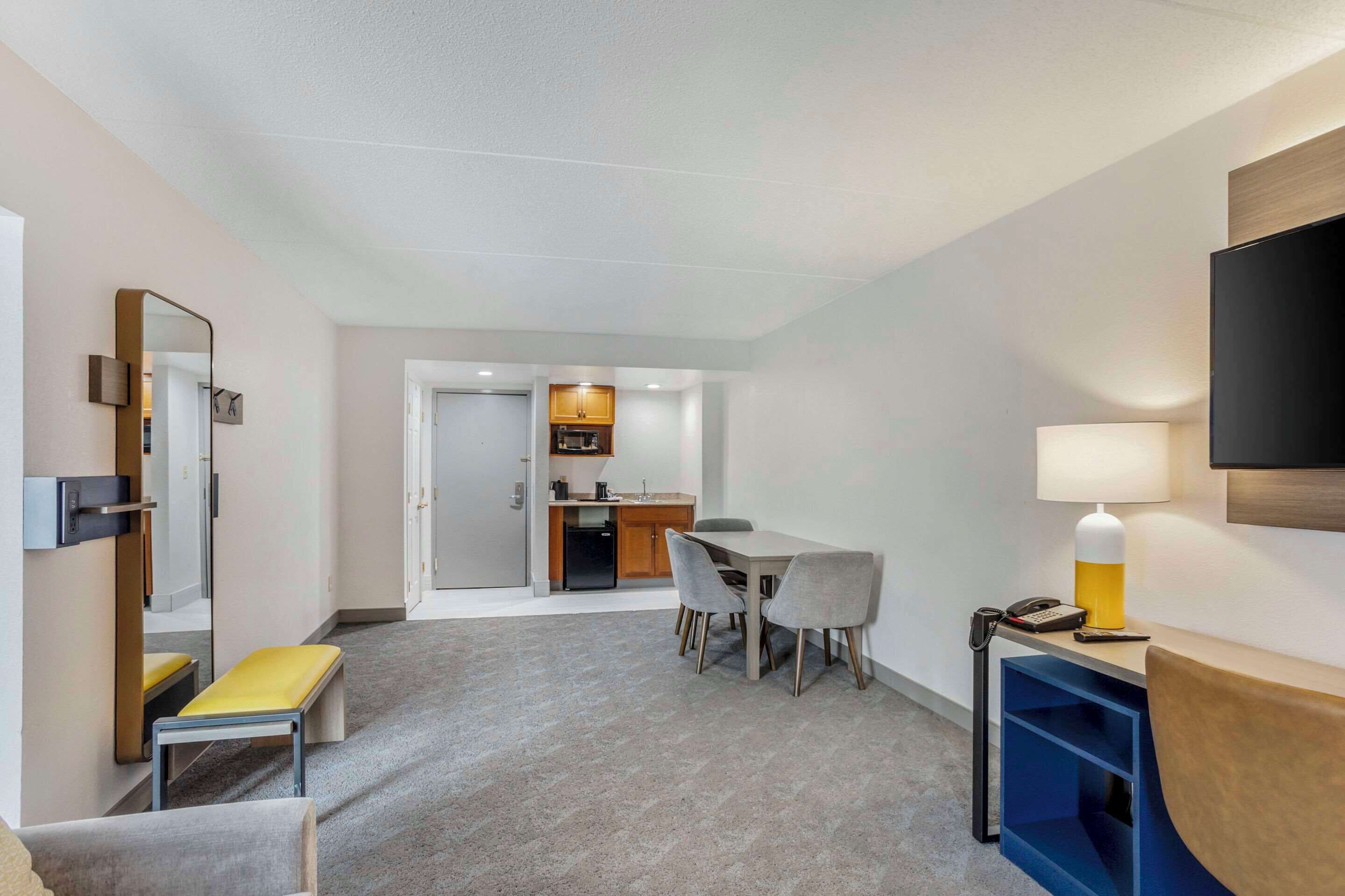A hotel room with a dining area, a small kitchenette, a bench, a TV, and a lamp.
