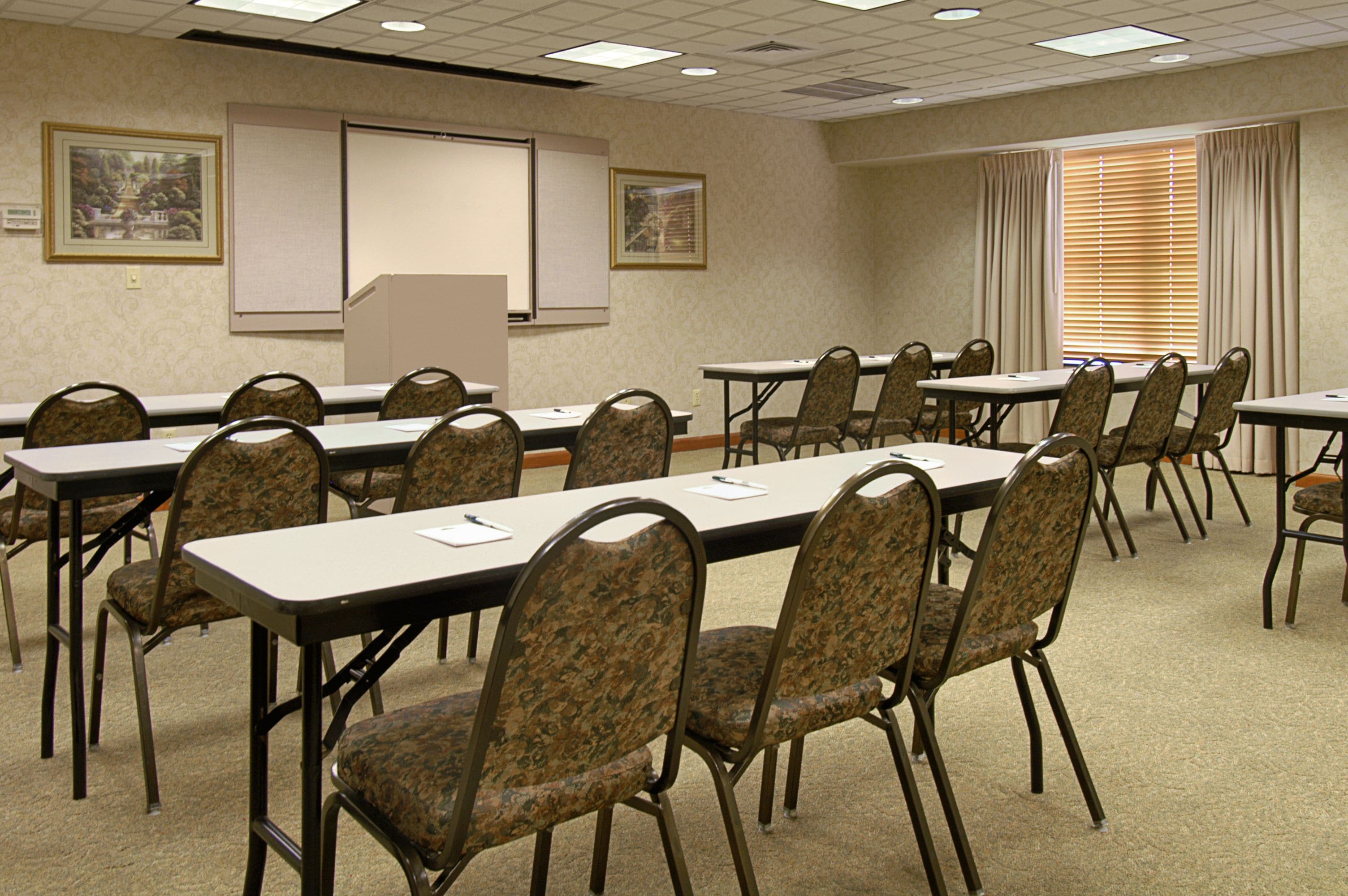 A conference room with long tables and patterned chairs, a projector screen, and framed pictures on the wall.