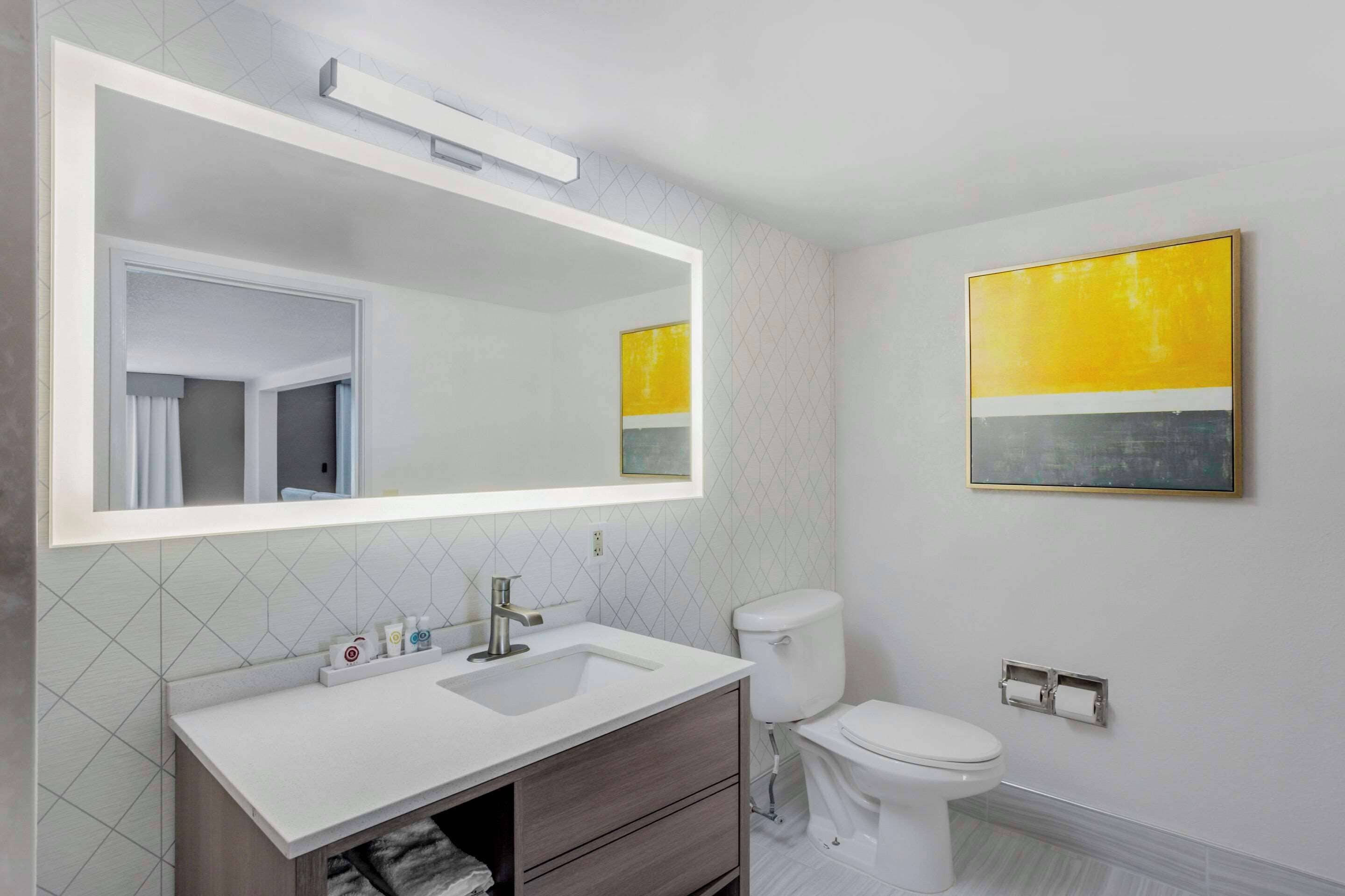 A bathroom with a white sink, a mirror, a toilet, and a yellow artwork on the wall.