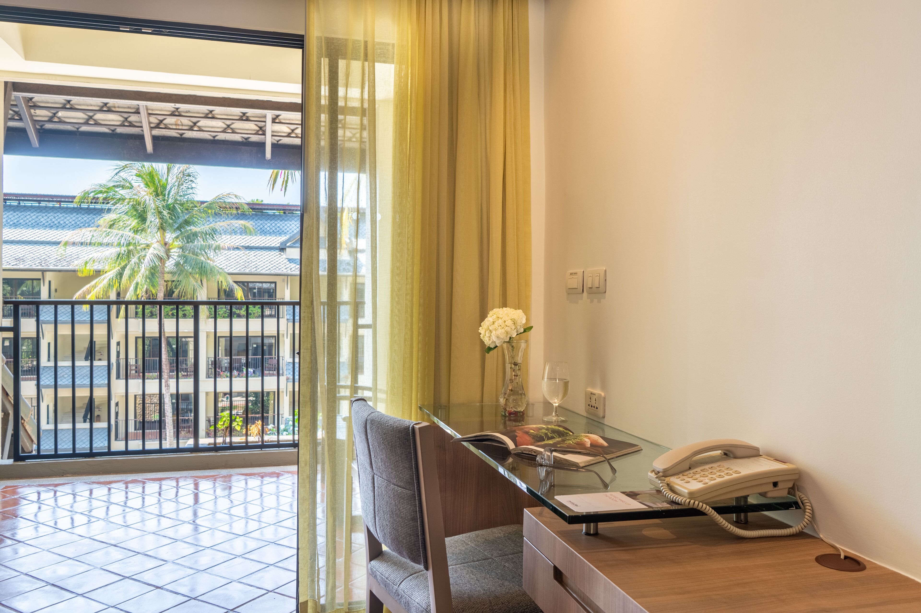 A hotel room with a balcony, a glass table with a telephone, a chair, and a vase with flowers.