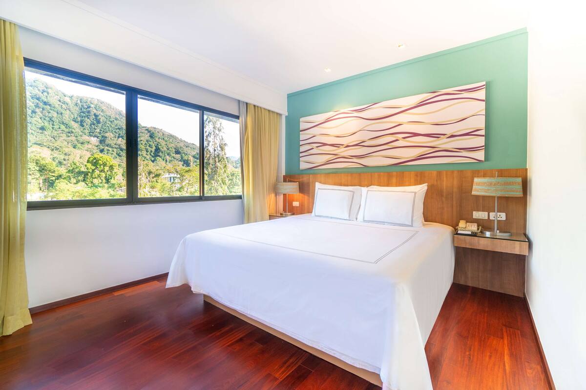 Une chambre d’hôtel avec un grand lit, une vue sur une colline boisée et une œuvre d’art moderne accrochée au mur.