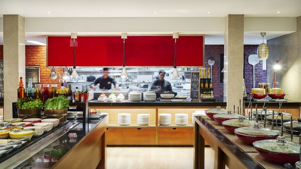 A buffet setup with various dishes and drinks, a kitchen area in the background, and a red partition. - Radisson Resort and Suites Phuket (Kamala)