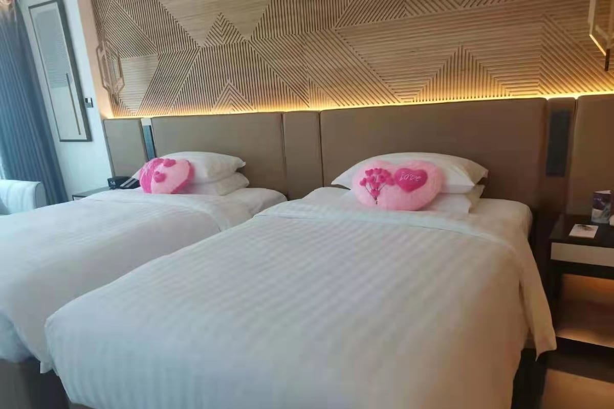 Ein Hotelzimmer mit zwei Betten, jeweils mit weißer Bettdecke und einem dekorativen, herzförmigen Kissen in Rosa.